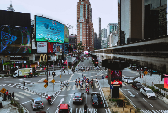 Busy urban intersection with pedestrians crossing, traffic signals, and prominent billboards, illustrating complex driving conditions relevant for road test preparation.
