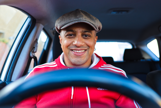 Smiling man in a red jacket and cap sitting behind the steering wheel of a car, representing confidence and readiness for a driving test in New York.