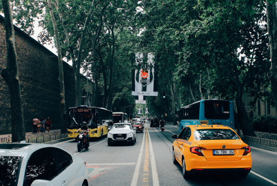 Busy city street with various vehicles, including a yellow taxi and a white car, surrounded by trees and a large banner overhead, representing urban driving conditions relevant for road test preparation.