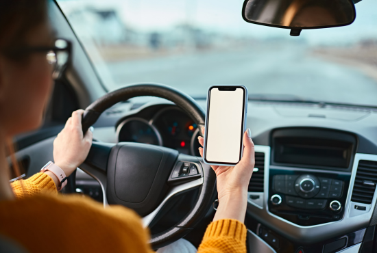 Person driving a car holding a smartphone, emphasizing safe driving practices and preparation for the New York State Road Test.
